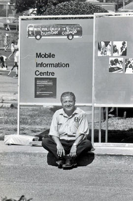 Photograph of Joe Miceli Sitting In Front of a Mobile Information Centre Sign