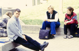 Photograph of students sitting on the curb in the courtyard behind building "E"