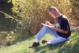 Photograph of a student sitting by the pond in the arboretum writing in a journal.