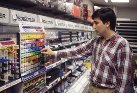 A photograph of a student browsing a wall of writing utensils in the Bookstore