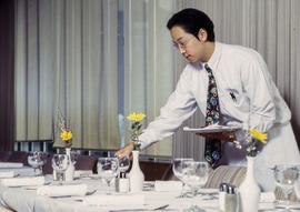Photograph of a hospitality student dressed in uniform sets the table in the Humber Room