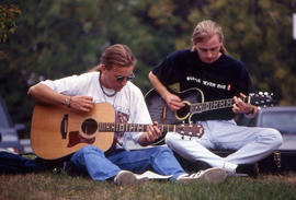 Photograph of two students sitting on the lawn, strumming on their guitars